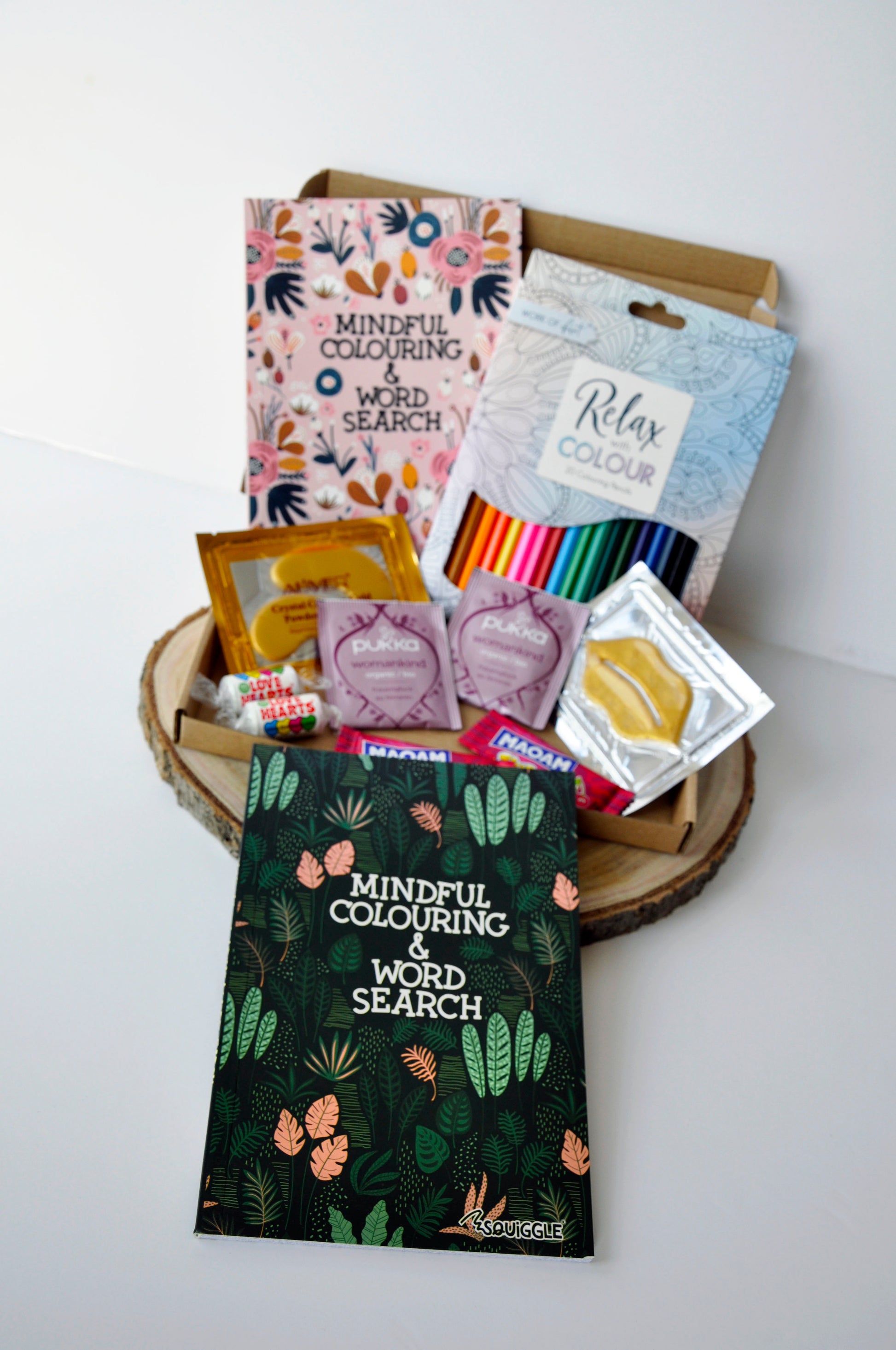 medium lettebox box showing both options of mindful coliuring and word search books, one in green and one in pink, with 2 pukka womenkind teas. 2 love heart sweets, 2 maoam sweets, gold collagen eye mask, lip mask